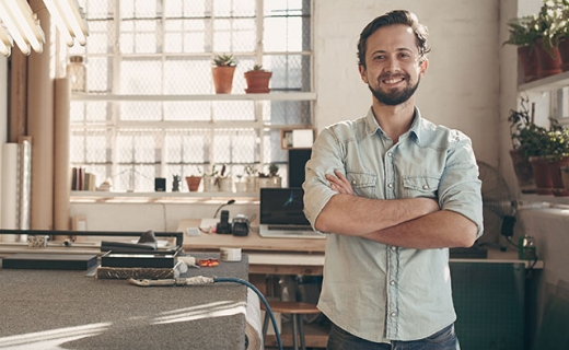 Person mit verschränkten Armen steht in einem hellen Studio oder einer Werkstatt mit Werkzeugen, Pflanzen und großem Fenster.